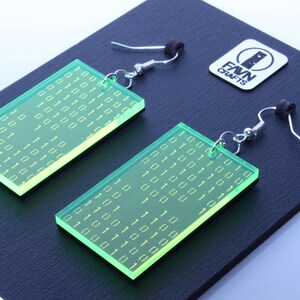 K&ouml;nnte beinhalten: Ein Paar rechteckige, neongr&uuml;ne Ohrringe mit einem Bin&auml;rcode-Muster. Die Ohrringe h&auml;ngen an silberfarbenen Haken und schwarzen Gummistopfen. Ein wei&szlig;es Logo mit dem Text "FAVN CRAFTS" ist ebenfalls sichtbar.