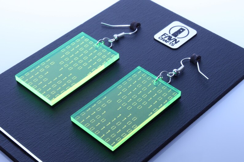 K&ouml;nnte beinhalten: Ein Paar rechteckige, neongr&uuml;ne Ohrringe mit einem Bin&auml;rcode-Muster. Die Ohrringe h&auml;ngen an silberfarbenen Haken und schwarzen Gummistopfen. Ein wei&szlig;es Logo mit dem Text "FAVN CRAFTS" ist ebenfalls sichtbar.