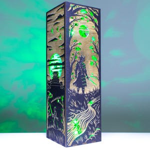 May include: A tall, rectangular wooden lamp featuring intricate laser-cut panels that depict a samurai standing in a forest landscape with a river and a large moon. The lamp emits a vibrant green light through the cutouts, highlighting the dark wood.