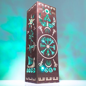 Peut inclure: Une lampe rectangulaire haute en bois brun foncé avec des motifs complexes découpés au laser, incluant un symbole Vegvisir central, un navire viking et divers motifs runiques. Les découpes sont éclairées par une lumière sarcelle vive.