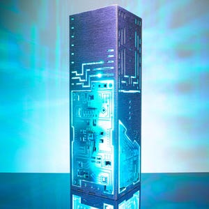 May include: A tall, rectangular tower lamp with a dark, textured surface featuring intricate, laser-cut circuit board patterns. The lamp emits a vibrant, glowing cyan light from within, illuminating the geometric pathways and electronic traces.