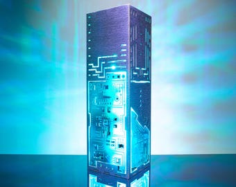 Peut inclure: Une lampe tour rectangulaire et haute avec une surface sombre et texturée présentant des motifs complexes de circuits imprimés découpés au laser. La lampe émet une lumière cyan vive et rayonnante, illuminant les tracés électroniques.