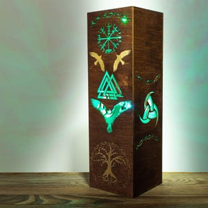 Pode incluir: Uma luminária de madeira alta e retangular com acabamento castanho escuro. O painel frontal apresenta desenhos cortados a laser, incluindo um Vegvisir, um Valknut e uma árvore, iluminados por dentro por uma luz verde brilhante através dos recortes.