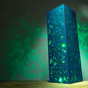 May include: A blue wooden lamp with a cutout design of sea turtles and seaweed. The lamp is lit and casting a green glow on the wall behind it.