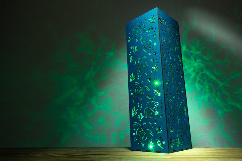 May include: A blue wooden lamp with a cutout design of sea turtles and seaweed. The lamp is lit and casting a green glow on the wall behind it.