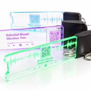 Op de afbeelding: Drie rechthoekige acryl sleutelhangers met zwarte plastic doppen en metalen ringen. Elke sleutelhanger heeft een gekleurd lichteffect, een QR-code, een geluidsgolfgrafiek en muziekspeler-pictogrammen. Tekst bevat Paper Lanterns, Celestial Roads en Dream Circuit.