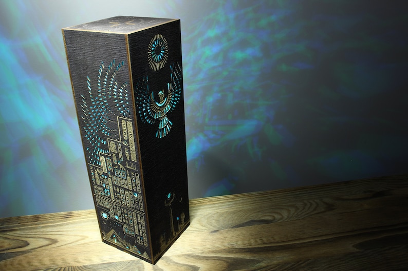 May include: A black wooden lamp with a gold and blue design of an Egyptian-style bird and hieroglyphics. The lamp is lit and casting a blue glow.