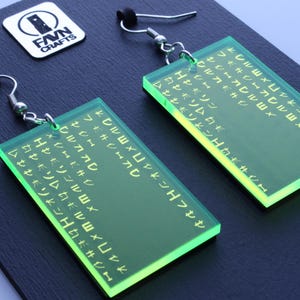 May include: A pair of neon green rectangular earrings with etched text. The earrings hang from silver-coloured hooks and black rubber stoppers. The earrings are displayed on a dark blue surface, with a white and black logo in the background.