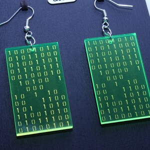K&ouml;nnte beinhalten: Ein Paar rechteckige Ohrringe in Neongr&uuml;n. Jeder Ohrring zeigt eine Reihe von Bin&auml;rcode-Zahlen in Wei&szlig;. Die Ohrringe h&auml;ngen an silberfarbenen Fischhaken-Ohrdr&auml;hten. Die Ohrringe werden auf einer schwarzen Oberfl&auml;che pr&auml;sentiert.