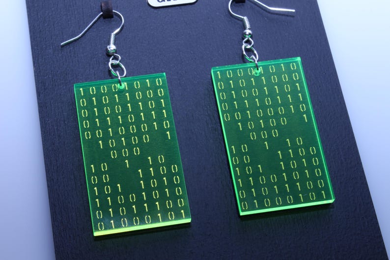 K&ouml;nnte beinhalten: Ein Paar rechteckige Ohrringe in Neongr&uuml;n. Jeder Ohrring zeigt eine Reihe von Bin&auml;rcode-Zahlen in Wei&szlig;. Die Ohrringe h&auml;ngen an silberfarbenen Fischhaken-Ohrdr&auml;hten. Die Ohrringe werden auf einer schwarzen Oberfl&auml;che pr&auml;sentiert.
