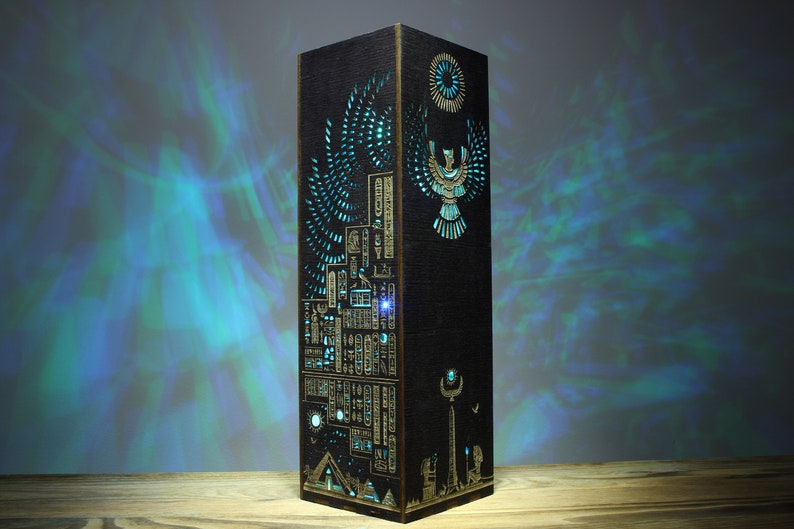 May include: A black wooden lamp with intricate laser cut designs depicting Egyptian hieroglyphs, a winged sun god, and a pyramid. The lamp is illuminated with a blue light, casting a shadow on the wall behind it.