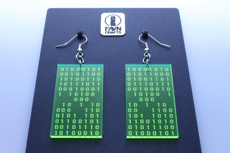 Pu&ograve; includere: Un paio di orecchini rettangolari verde neon con motivi a codice binario. Ogni orecchino ha un gancio argentato e una serie di numeri in una griglia. Gli orecchini sono esposti su una superficie nera con un logo.