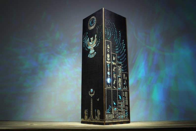 May include: A black wooden lamp with a cutout design of an Egyptian-style winged figure, hieroglyphics, and a sun disc. The lamp is illuminated with a blue light, casting a pattern on the wall behind it.