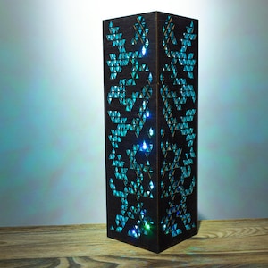 May include: A tall, rectangular dark brown wooden lamp with intricate triangular cutouts. The lamp emits a bright, glowing turquoise light through the geometric patterns, casting a soft blue illumination on the surrounding area.