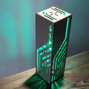 May include: A tall, rectangular wooden lamp with a dark finish and intricate, laser-cut geometric patterns on its sides. The lamp emits a bright green light from within, casting a glow through the cutouts and onto the surrounding surface.