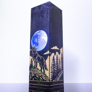 Könnte beinhalten: Eine hohe, rechteckige, dunkel gefärbte Lampe mit einem lasergeschnittenen Design eines Wolfes, der einen hellen, beleuchteten Vollmond anheult. Die Basis der Szene zeigt einen Kiefernwald und felsiges Gelände mit grünen Akzenten, und die oberen Kanten weisen komplizierte keltische Knotenmuster auf.