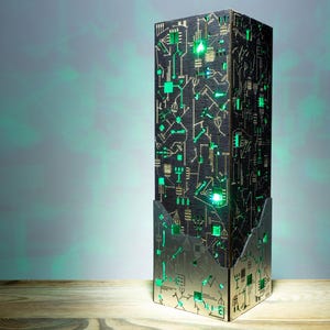 Peut inclure: Une lampe rectangulaire haute avec une surface noire texturée ornée de motifs de circuits imprimés dorés et de découpes. La lampe émet une lumière verte vive de l'intérieur, illuminant les motifs géométriques et la base métallique.