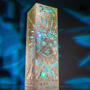 May include: A tall, rectangular, gold-toned lamp with a laser-cut design featuring a central face surrounded by radiating sunburst lines. The cutouts emit a bright, vibrant cyan light, casting blue patterns onto the surrounding dark space.