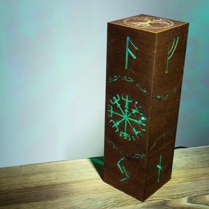 Könnte beinhalten: Eine hohe, rechteckige Holzlampe mit dunkelbraunem Finish. Die Seiten zeigen ausgeschnittene nordische Runen und ein Vegvisir-Symbol, das von hellem grünem Licht beleuchtet wird. Die Oberseite zeigt ein eingraviertes Lebensbaum-Motiv.