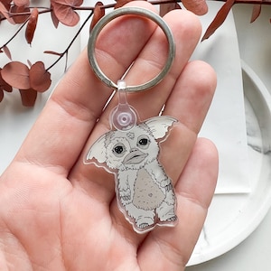 May include: A clear acrylic keychain featuring a cartoon illustration of a white furry creature with large eyes and a small mouth. The creature is standing on two legs and has large ears.