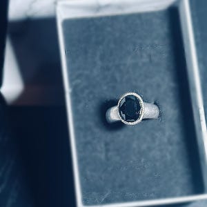 Puede incluir: Un anillo de plata con una gran piedra negra de forma ovalada, rodeada por un halo de piedras más pequeñas y transparentes. El anillo está engastado en una banda texturizada y se muestra en una caja blanca con interior azul oscuro.