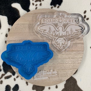 Peut inclure: Un moule en silicone bleu et un moule en plastique transparent d'un aigle avec les mots "Roam Free" et "Desert Savior" à l'intérieur des ailes. Les moules sont sur une surface en bois.