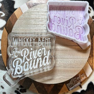 May include: Clear acrylic stamp and a purple silicone mold with the text "Whiskey Bent & River Bound". The stamp is layered with a clear handle. The mold has the text in reverse. A small clear stamp is also visible.