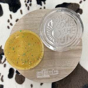 Può includere: Due sottobicchieri rotondi, uno trasparente con il testo "Here Comes the Sun" e l'altro giallo con glitter e un design a forma di sole. I sottobicchieri sono su una superficie di legno con il testo "sweet tea and me creations" stampato su di essa.