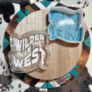 Può includere: Uno stampo rettangolare trasparente e uno stampo blu con la scritta "Wilder Than The West" su una superficie di legno con un bordo decorativo di forme geometriche turchesi, marroni e bianche. Lo stampo trasparente ha testo bianco.