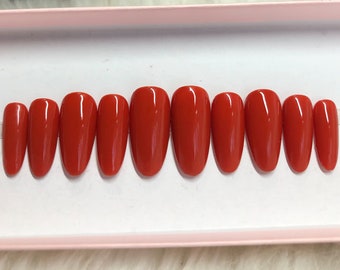 以下が含まれることがあります： 白い箱の中に、10本の赤い楕円形の付け爪が入っています。爪は、短いものから長いものまで、一列に並んでいます。