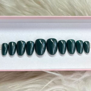May include: A set of ten dark green, oval-shaped press-on nails arranged in a row. The nails are displayed in a pink and white box. The nails are a solid color with a glossy finish. The nails are of varying sizes.
