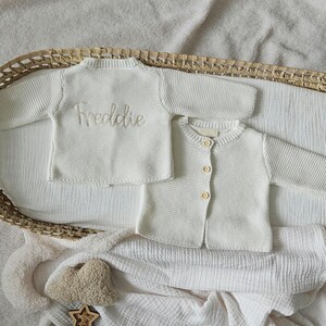 May include: Two white knitted baby cardigans, one with the name "Freddie" embroidered on the front. The cardigans are laid out on a white fabric background.