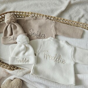 May include: Two knitted baby sweaters with pom-pom hats. One sweater is beige with the name "Sofia" embroidered on it. The other sweater is white with the name "Freddie" embroidered on it.