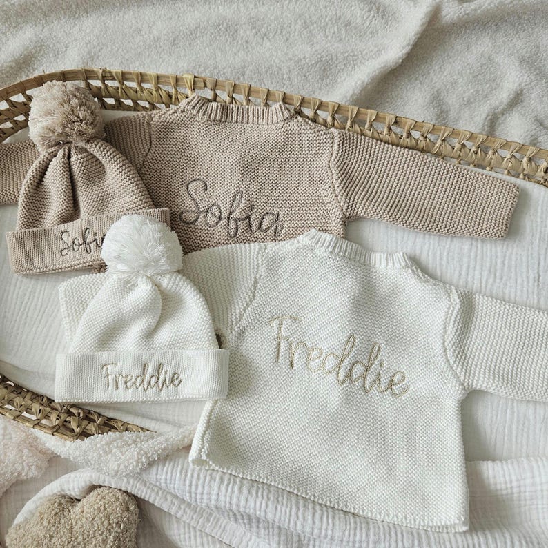 May include: Two knitted baby sweaters with pom-pom hats. One sweater is beige with the name "Sofia" embroidered on it. The other sweater is white with the name "Freddie" embroidered on it.
