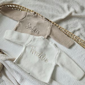May include: Two knitted baby sweaters, one beige and one white, with the names "Sofia" and "Freddie" embroidered on them.