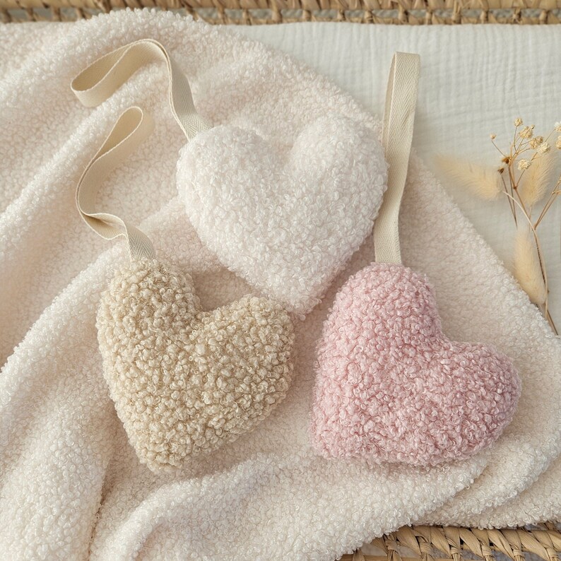May include: Three plush heart-shaped toys in white, beige, and pink. The toys are made of soft, fuzzy fabric and are hanging from a white blanket.