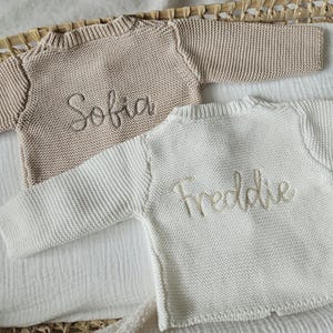 May include: Two knitted baby sweaters, one beige and one white, with the names "Sofia" and "Freddie" embroidered on them.