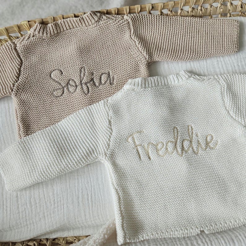 May include: Two knitted baby sweaters, one beige and one white, with the names "Sofia" and "Freddie" embroidered on them.