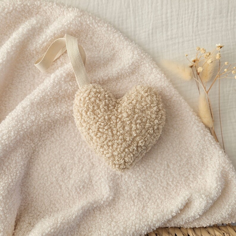 May include: A cream-colored heart-shaped plush toy with a fluffy texture. The toy is hanging from a white string and is resting on a white fuzzy surface.