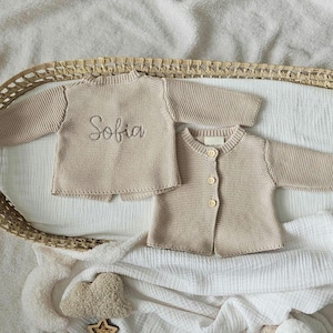 May include: Two beige knitted baby cardigans, one with the name "Sofia" embroidered on the back.