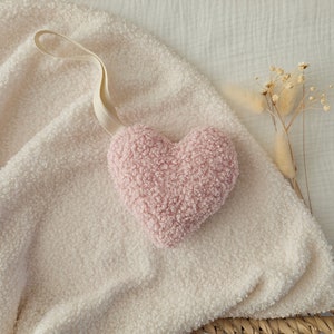 May include: A pink heart-shaped plush toy with a fluffy texture, hanging from a white string on a white fuzzy blanket.