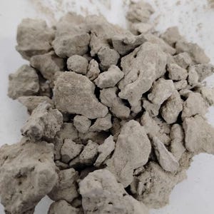 May include: A pile of irregularly shaped, light gray rocks. The rocks have a rough, porous texture and vary in size. The background is a plain white surface, providing contrast to the rocks. The overall appearance suggests a natural, earthy material.