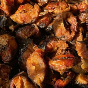 May include: Close-up of a pile of smoked fish fillets. The fish has a dark brown, almost black exterior with a reddish-brown interior. The texture appears flaky and slightly oily, with visible bones and fins. The image is well-lit, highlighting the details of the fish.