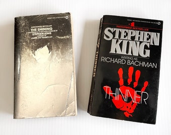 May include: Two paperback books, one with a white cover and the title "The Shining" by Stephen King, and the other with a black cover and the title "Thinner" by Stephen King writing as Richard Bachman.