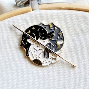 May include: A gold-toned metal pin featuring two black and white rabbits intertwined in a yin-yang design. The rabbits are surrounded by stars, a crescent moon, and a sun. The pin is on a white fabric background.