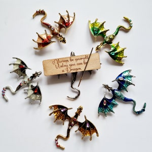 May include: Six colorful dragon brooches with jeweled accents are arranged around a wooden sign that reads "Always be yourself unless you can be a dragon."