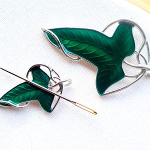 May include: Two silver-toned leaf-shaped brooches with vibrant green enamel and a needle. The leaves have detailed veining and are set in a decorative silver frame. The needle is gold-toned with a sharp point.