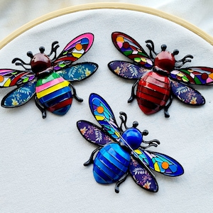 May include: Three colorful bee brooches with intricate details. The bees are made of metal and have a glossy finish. The first bee is red, yellow, and blue with floral patterns on its wings. The second bee is red, black, and white with floral patterns on its wings. The third bee is blue, purple, and yellow with floral patterns on its wings.