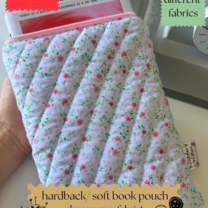 May include: A quilted fabric book pouch featuring a white background with a small floral pattern of pink flowers and green leaves. The pouch has a light pink zip closure and a small fabric tag on the side that reads Stitches by Ethan.