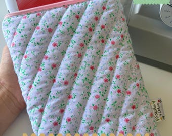 Op de afbeelding: Een gewatteerd stoffen boekenhoesje met een witte achtergrond en een klein bloemenpatroon van roze bloemen en groene blaadjes. Het hoesje heeft een lichtroze ritssluiting en een klein stoffen label aan de zijkant.